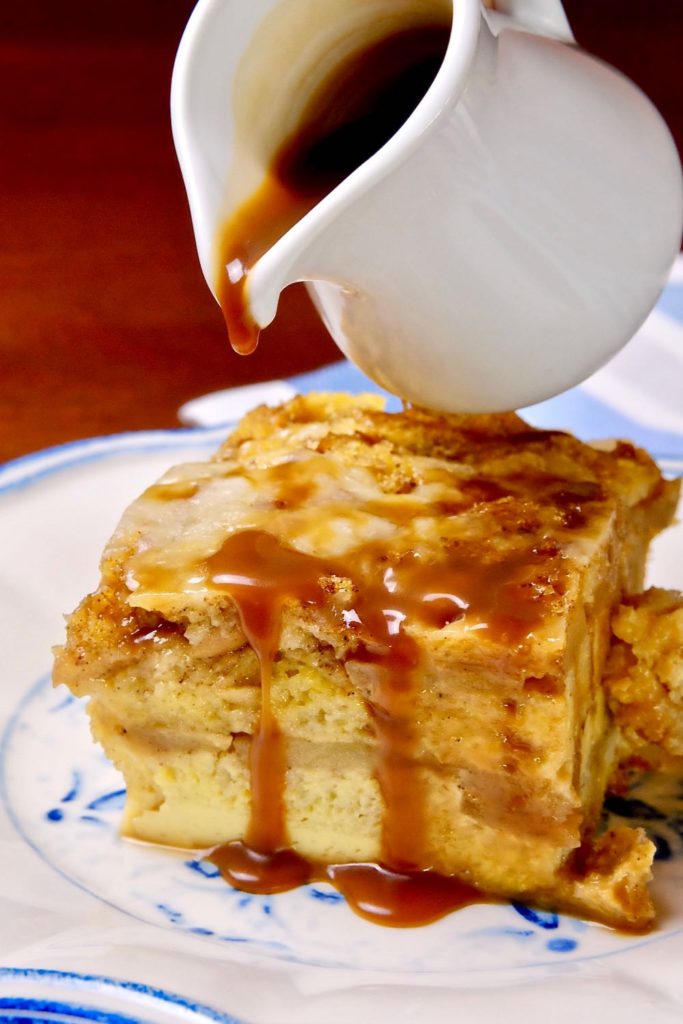 Apple Cinnamon Strata being drizzled with Apple Cider Caramel Sauce on white and blue plate with blue and white striped linen in background.