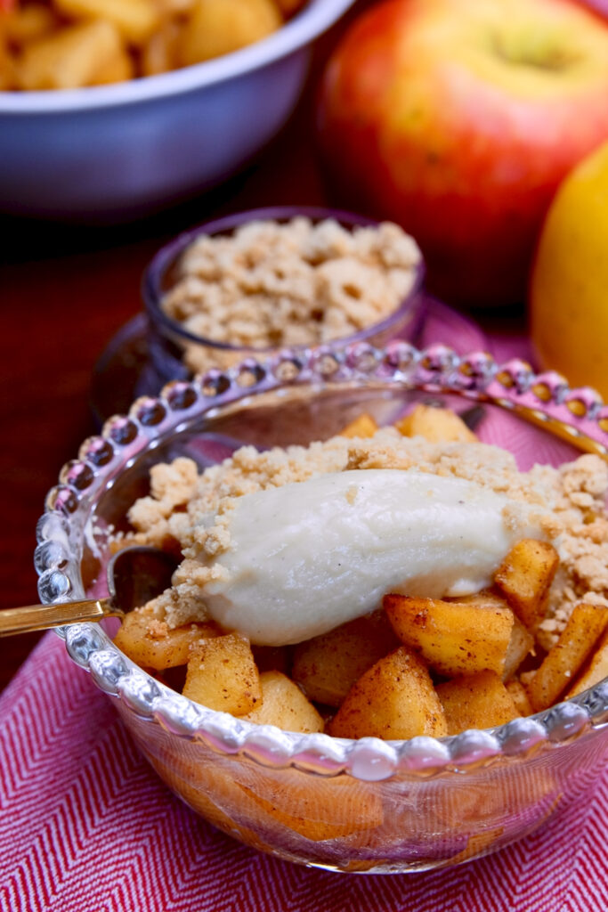 Apple and vanilla pudding parfait served in a glass bubble dessert bowl garnished with a cookie garnish is set on a red and white linen napkin.  Bowls of sauteed cinnamon apples and cookie crumbs are in left hand background with two fresh apples in right hand background.