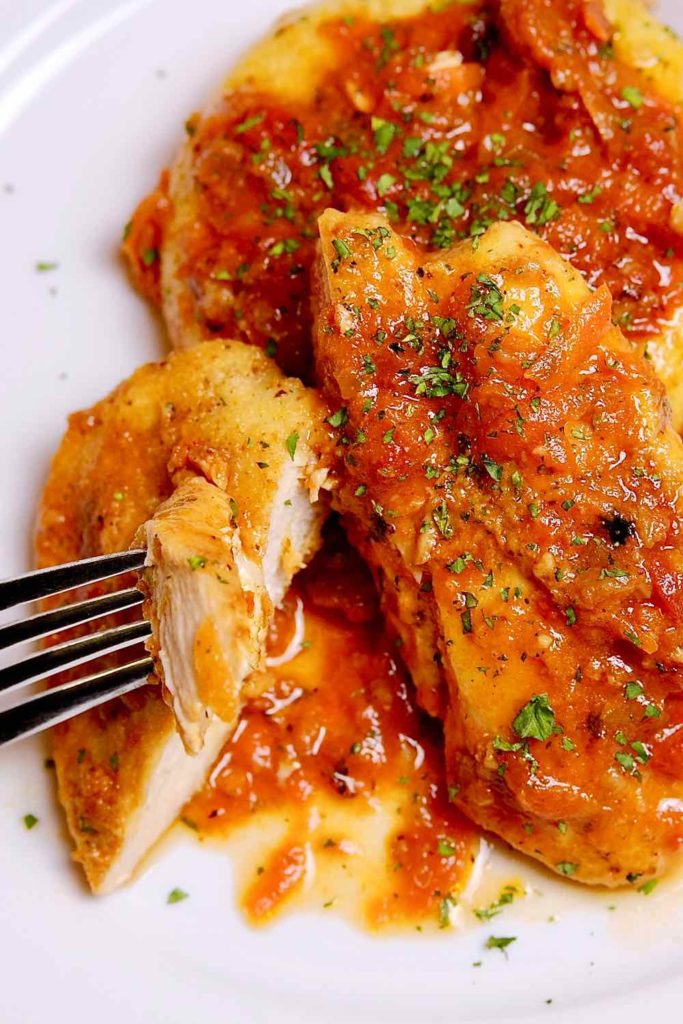 Chicken cacciatore served over polenta on a white plate  Bite shot of cut chicken on fork in the left hand side of dish. 