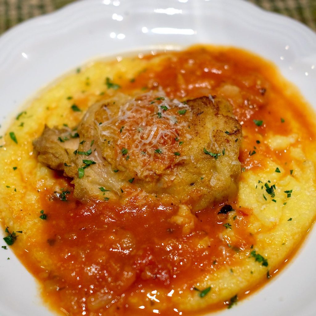 Chicken braised in marinara sauce and served over polenta in a white bowl garnished with chopped parsley.  