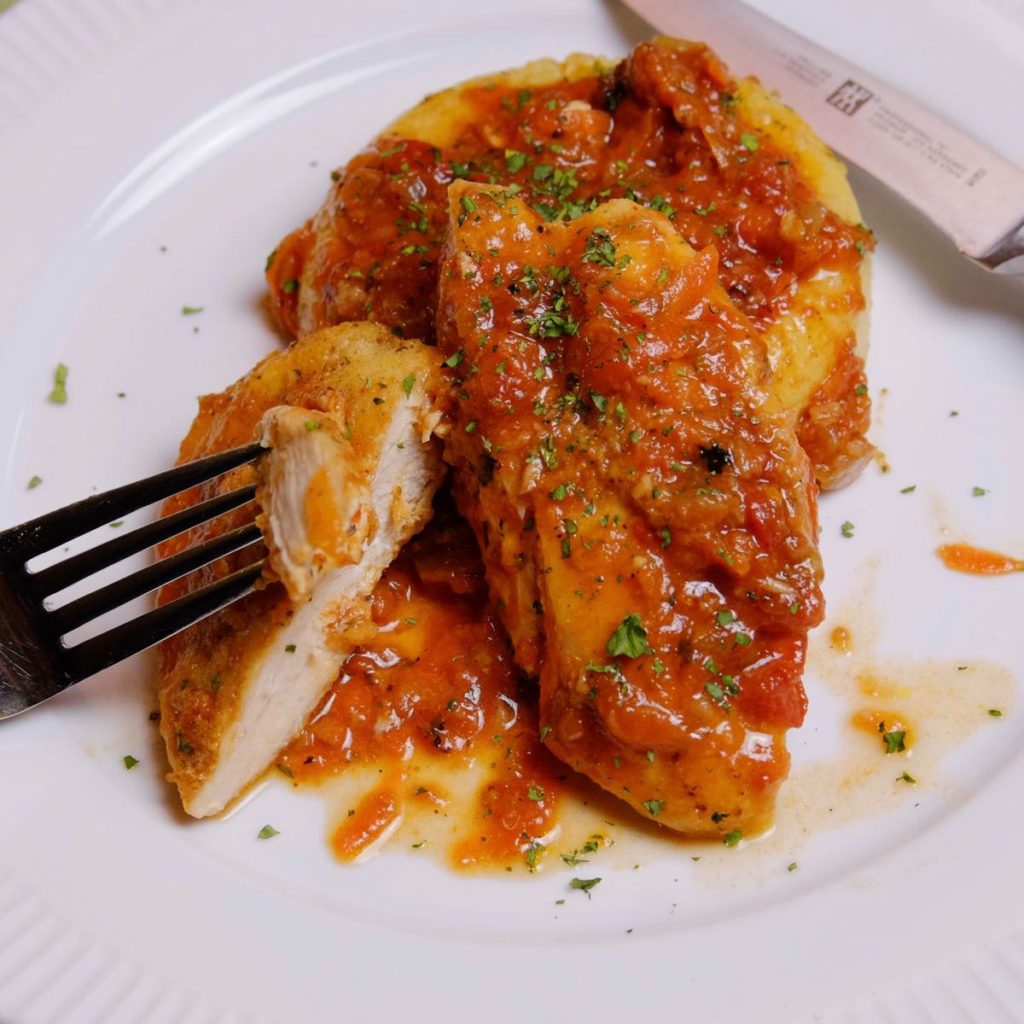 Chicken breast braised in a simple Cacciatore sauce on a white plate with polenta. Fork showing bite shot in foreground of plate. The chicken dish is garnished with fresh minced parsley and a knife is set on upper edge of plate.