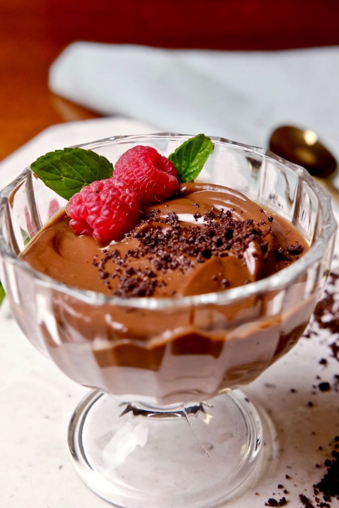Creamy chocolate pudding served in dessert glass is garnished ith chocolate sand, fresh mint and raspberries. Glass is set on a white marble surface with chocolate sand scattered in background. 
