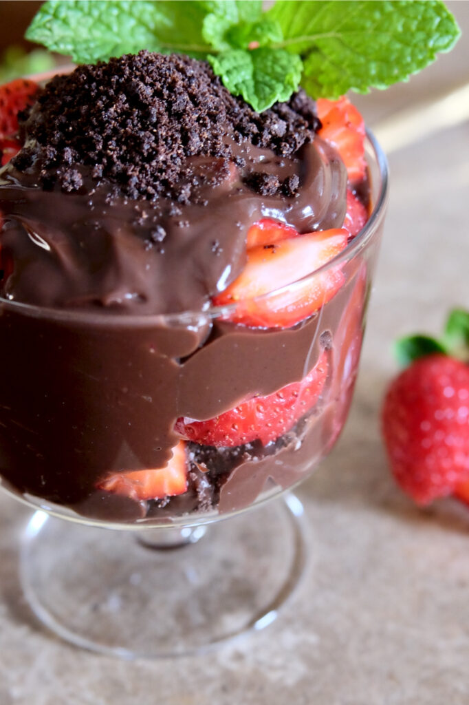 Chocolate pudding layered with sliced fresh strawberries and chocolate cookie crumbs in a glass dessert bowl and garnished with fresh mint.  Dessert bowl is set on beige granite with fresh strawberry lying in the fore front.