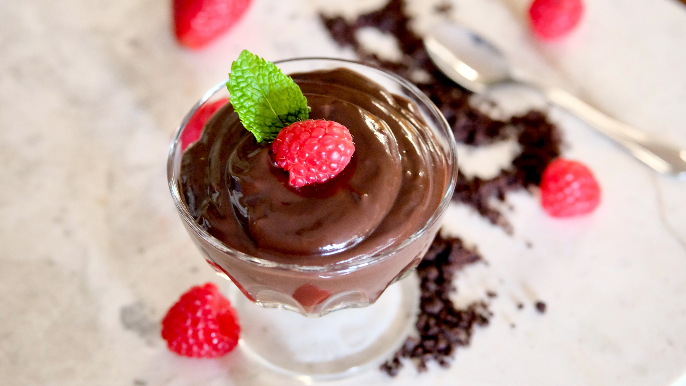 Chocolate Pudding in dessert glass garnished with raspberry and mint leave is sitting on a beige and white marble top. Spoon is in bakground of upper right with chocolate sand scattered about the base of dessert glass with fresh strawberries and raspberries.
