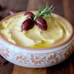 Hummus in decorative clay bowl garnished with rosemary, Kalamata olives and olive oil
