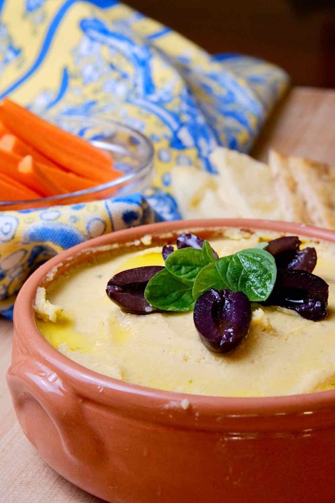 Hummus in a terracotta serving bowl garnished with fresh sprig of oregano and Kalamata olives. Slices of  Naan bread arranged around the bowl and a blue and yellow linen in background with a bowl of carrot sticks. 