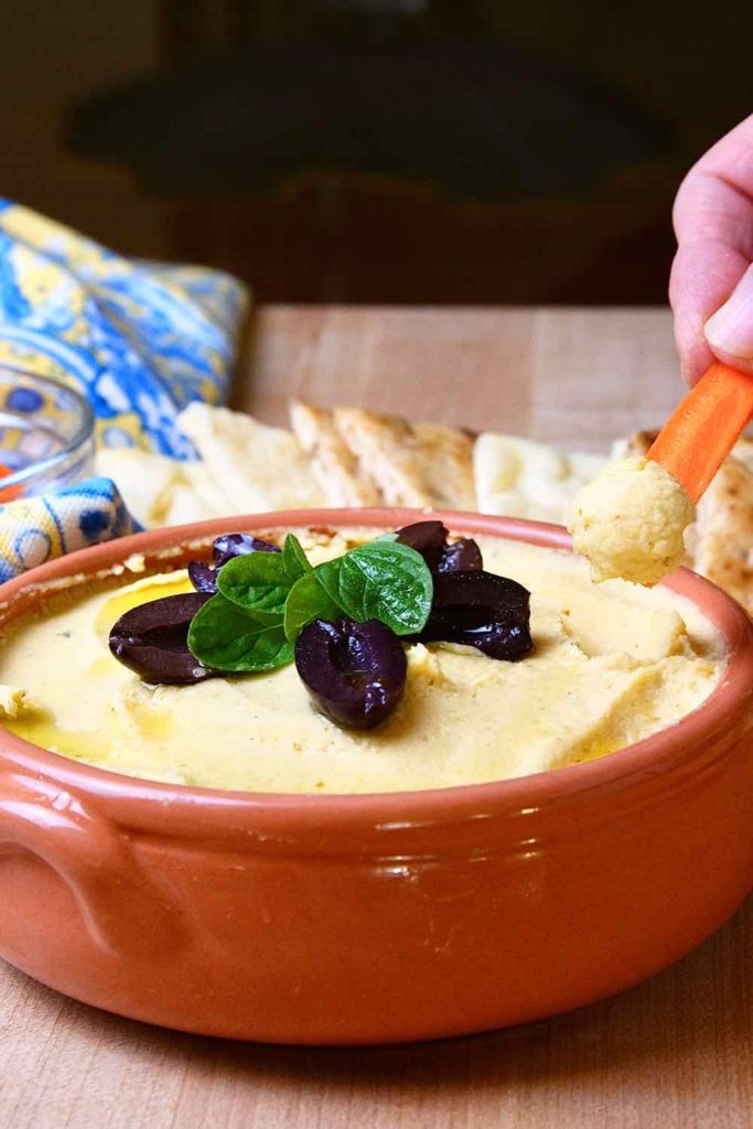 Hummus served in a terracotta serving bowl garnished with olive oil, Kalamata olives and fresh oregano sprig.  Bite shot of Carrot stick being scooped in bowl with cut slices of naan and blue and yellow linen in background. 