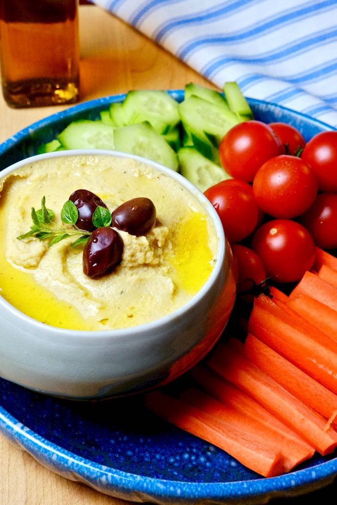 A bowl of hummus on a blue platter served with carrot sticks, tomatoes and cucumber slices.  Blue and white striped linen and olive oil in background.