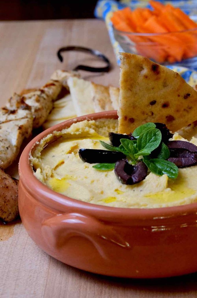 Hummus served in a terracotta serving bowl garnished with olive oil, kalamata olives and fresh oregano sprig.  Naan bread being scooped in bowl and chicken kebab and cut slices of naan in background. 