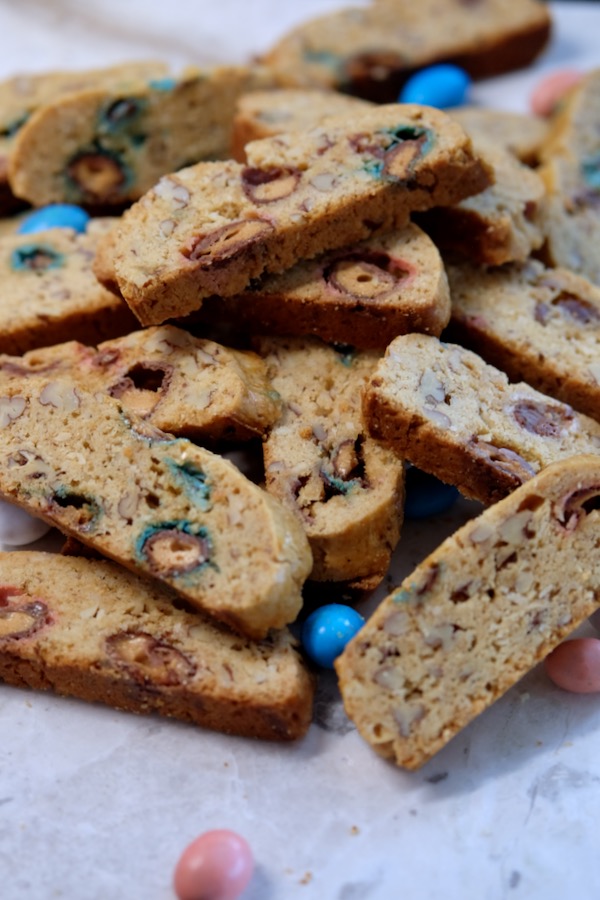 Italian Easter Biscotti are piled on top of a white marble board with blue, pink and yellow candy Robin Eggs scattered in the background and foreground.
