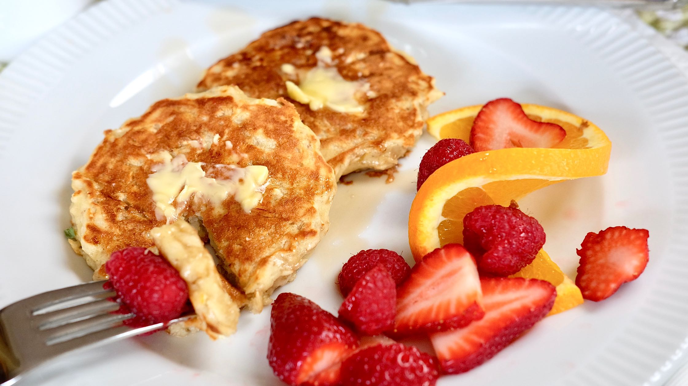 Lemon Breeze Apple Pancakes with butter and syrup on white plate with fresh strawberries and orange slice in background.