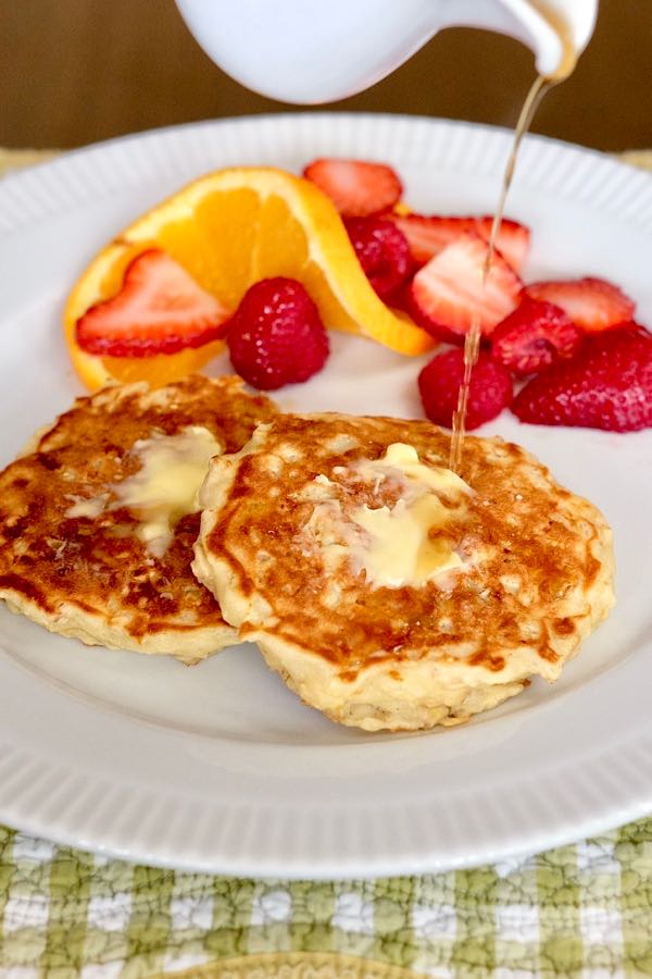 Lemon Breeze Apple Pancakes served with butter and syrup on a white plate, with fresh strawberries and orange slice.