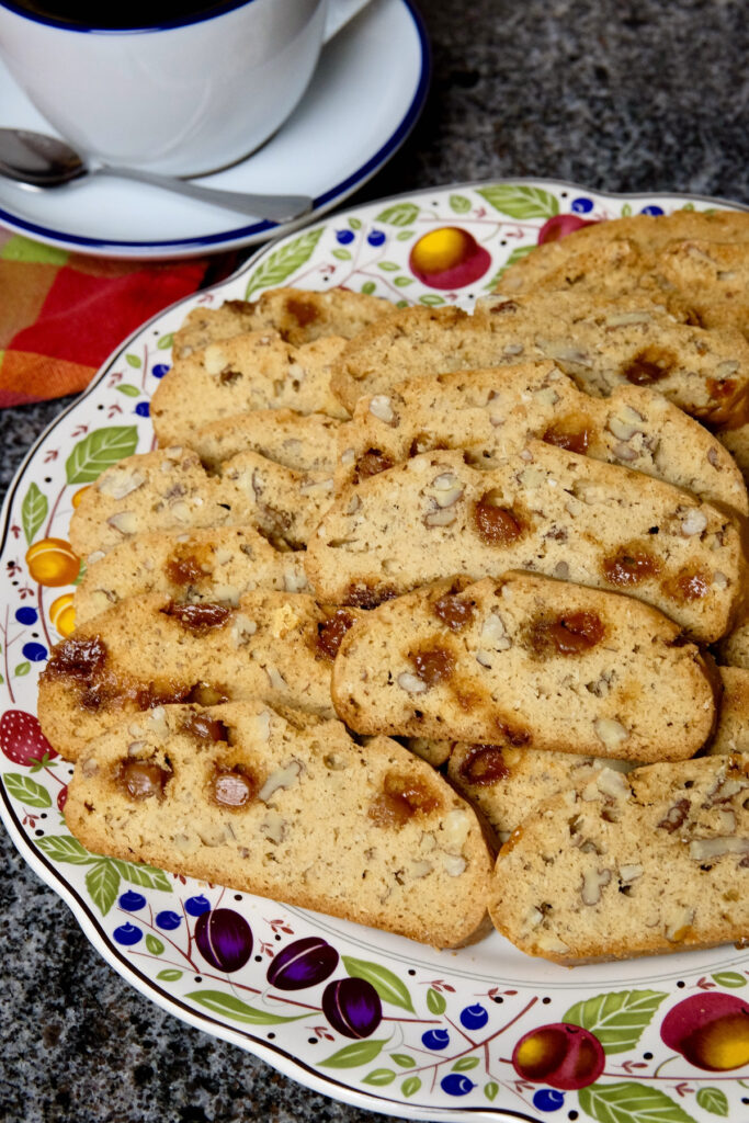 Slices of maple biscotti arranged on a multi-colored plate.  Cappuccino cup and saucer sitting on burgundy linen in background. cup