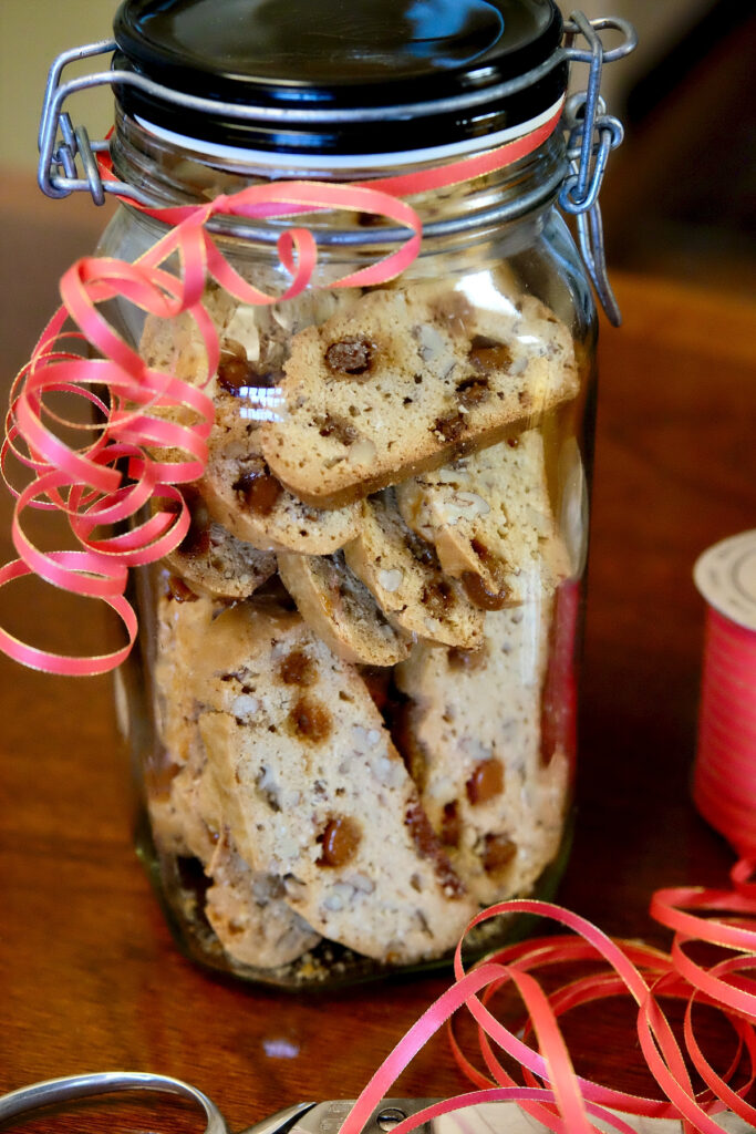 Maple Pecan Biscotti stacked in a large air tight jar tied with red holiday curling ribbon is sitting on a walnut table.  Spool of red ribbon and scissors are in front right foreground.