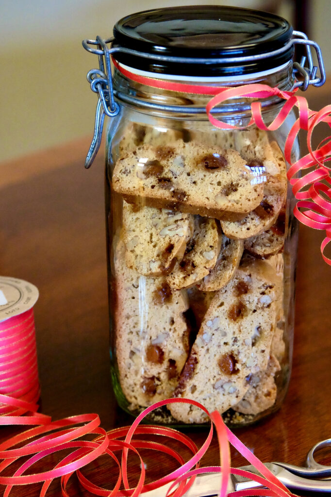 Maple Pecan biscotti packed in a glass container wrapped with red ribbon set on a walnut table.  A pair of scissors and spool of red curling ribbon are set in forefront.