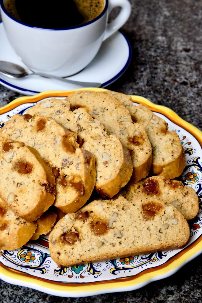 Maple Pecan Biscotti arranged on a multi-colored Italian plate set on black granite with coffee in a blue-rimmed white cup and saucer set in background.