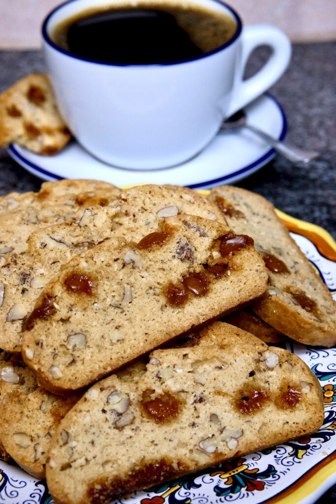 Maple pecan biscotti arranged on a multi-colored Italian plate.  Blue rimmed white coffee cup is set on a matching saucer with spoon and bitten biscotti in background.