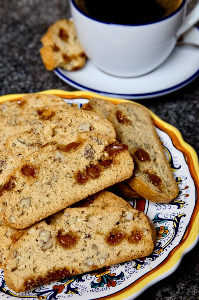 Maple pecan biscotti  served on an multi-colored Italian plate set on black marble.  White coffee cup filled with coffee is set on a blue rimmed white saucer with bitten biscotti set on edge is in bacground.