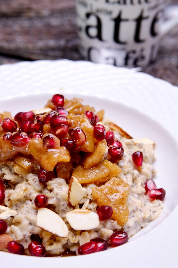 Oatmeal served in a white bowl topped with sauteed cinnamon apples, pomegranate arils and sliced almonds. Black and white latte cup in background>