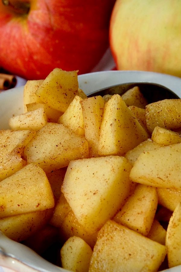 Sauteed Cinnamon Apples in beige serving bowl setting on a red and white plaid linen overlay with fresh apples and cinnamon sticks in background