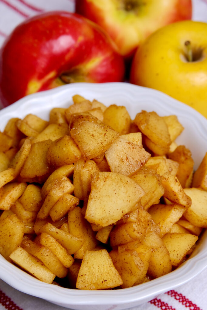 Sauteed cinnamon apples served in a white bowl set on a red striped white linen.  Three fresh apples are in background.