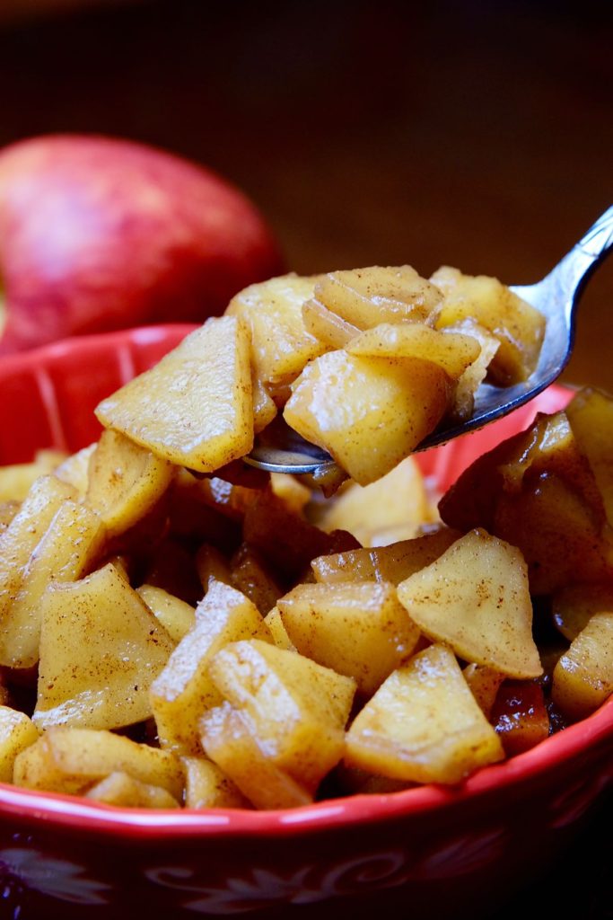 Sauteed Cinnamon Apples in a red bowl showing spoonful of apples. Background is dramatic darkness with apple set behind bowl.