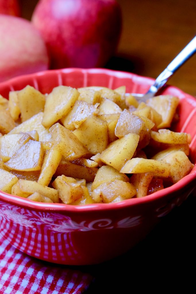 Red bowl filled with sauteed cinnamon apples set on a red and white gingham napkin. Two apples sit in the background on dark wood backdrop.