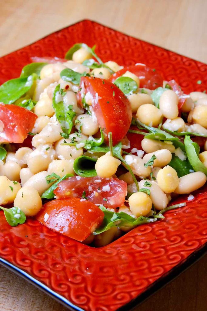 Sicilian Bean and Tomato Salad in a red square serving dish garnished with fresh arugula and grated Grana Padano. 