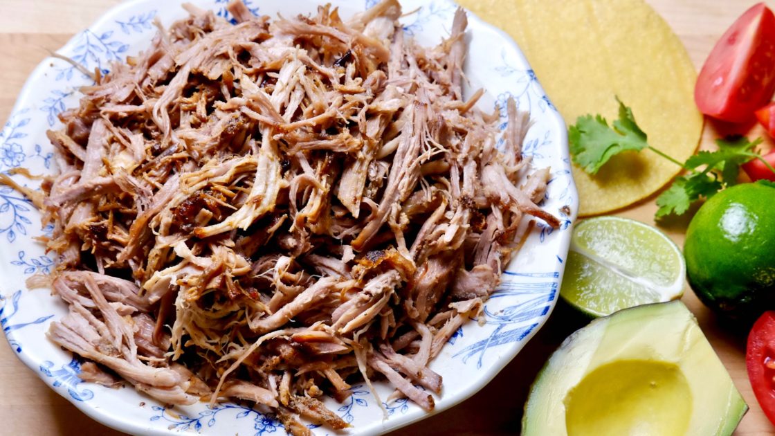 White and blue bowl filled with pork carnitas on maple board. Avocado, tomato and lime wedges in background with tortillas.