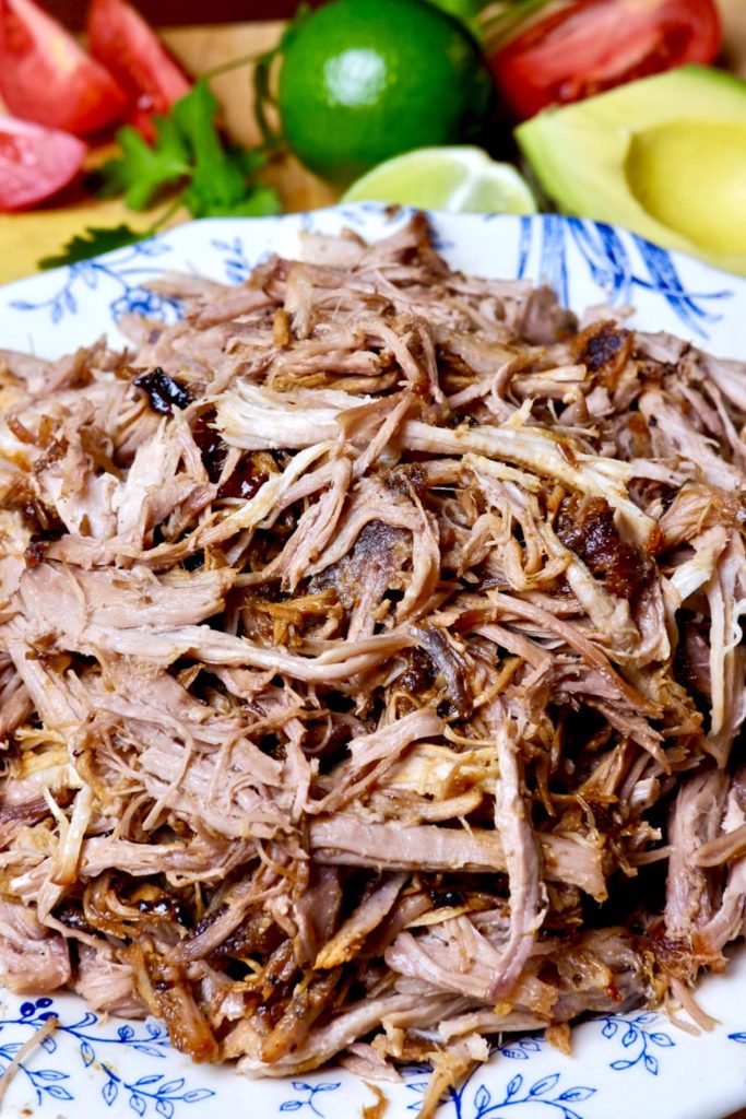 White and blue bowl piled high with shredded pork carnitas.  Tortillas, lime, tomato wedges and avocado in background.