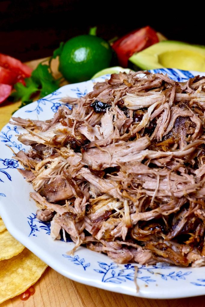 Slow roasted Carnitas shredded in a blue in white bowl with avocado slices, lime and tomato wedges in background.