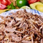 Roasted and shredded carnitas in white & blue bowl with lime and tomato wedges in background.