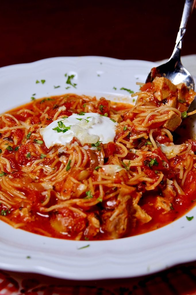 Sopa Seca de Fideo in white soup bowl garnished with a dollop of sour cream and freshly minced cilantro. 
 Spoon lifting from back right of bowl showing bite shot of soup.  