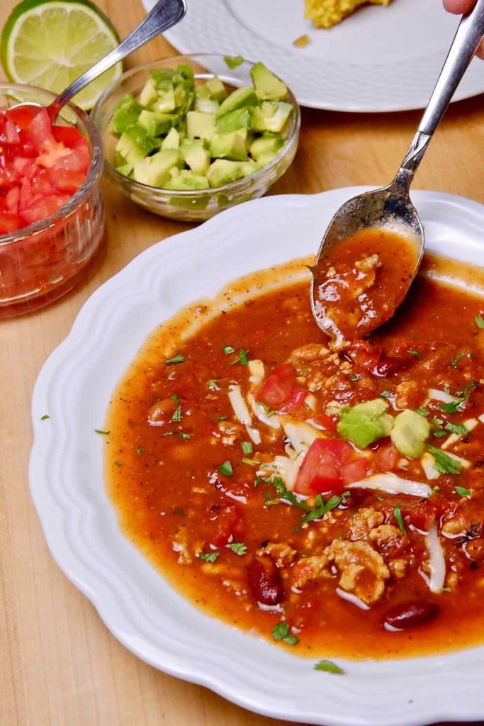 Turkey Chili Soup in white bowl with spoon shot of soup.  Containers of chopped tomato and avocado in background with slice of cornbread on white plate.