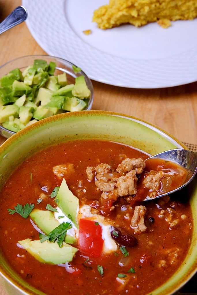 Bite shot showing soup garnished with sour cream, tomato and avocado served in a green soup bowl.  Chopped avocado in glass bowl in background. 