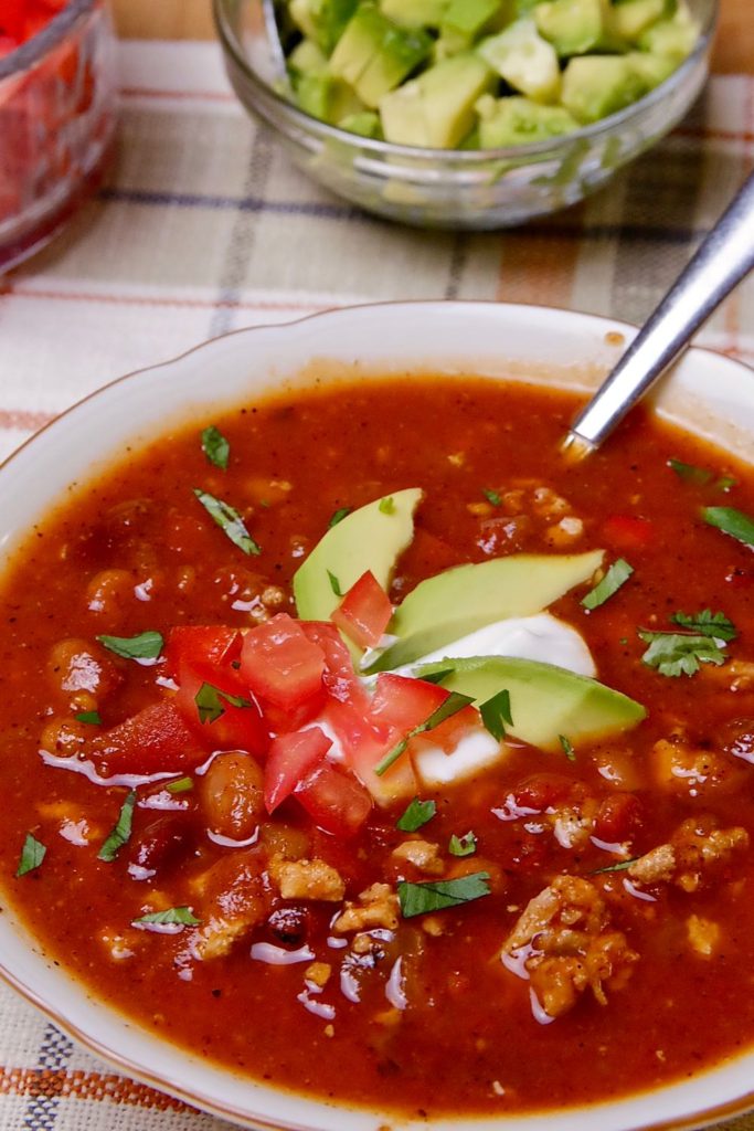 Turkey Chili Soup garnished with sour cream, avocado and tomato. Glass bowls with chopped tomato and avocado are set in background on a beige and brown linen placemat.