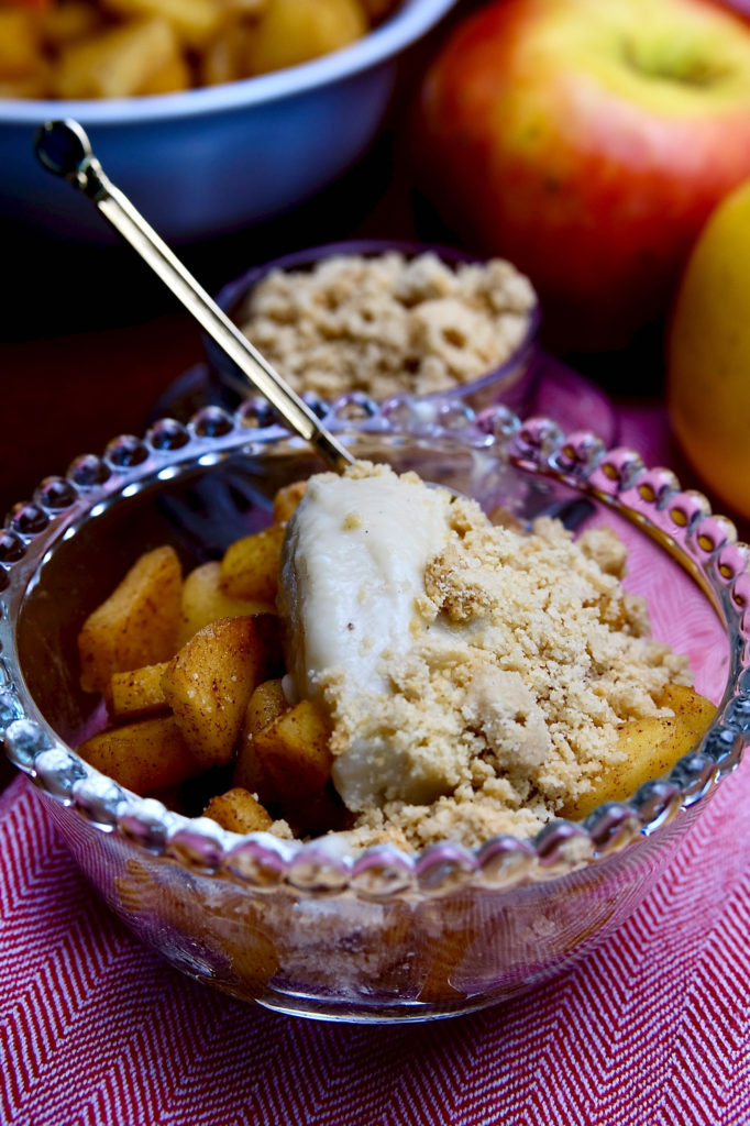 Sauteed cinnamon apples in a glass cup topped with vanilla pudding made from almond milk and sprinkled with edible sand.  The bowl is set on a light red linen and a gold spoon is setting in the back of the bowl.  A blue bowl of sauteed apples and a small glass bowl of edible sand is in background with two fresh apples.