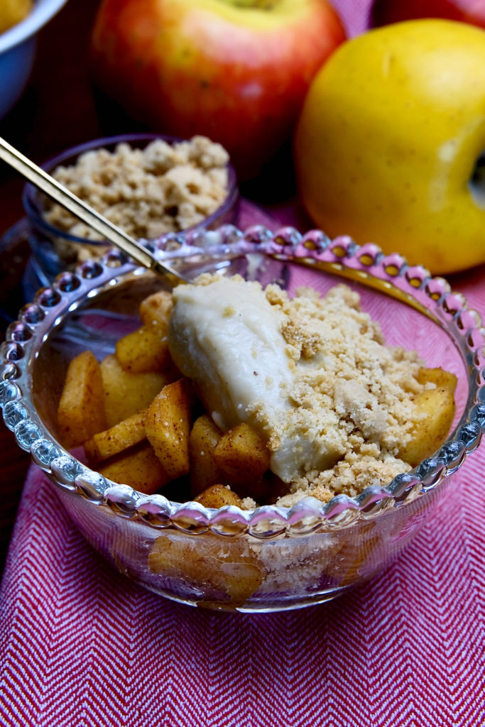 Sauteed cinnamon apples in a glass cup topped with vanilla pudding made from almond milk and sprinkled with edible sand. The bowl is set on a light red linen and a gold spoon is setting in the back of the bowl. A blue bowl of sauteed apples and a small glass bowl of edible sand is in background with two fresh apples.