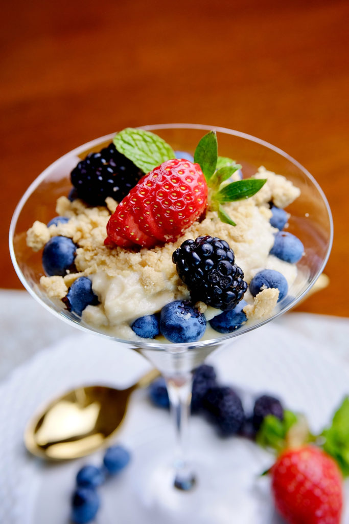 Dairy free vanilla pudding layered with fresh blueberries served in a martini glass.  The dessert is garnished with additional fresh blueberries, blackberries and a sliced strawberry then sprinkled with edible cookie sand.  Fresh berries are scattered around the base of the glass with a gold spoon.