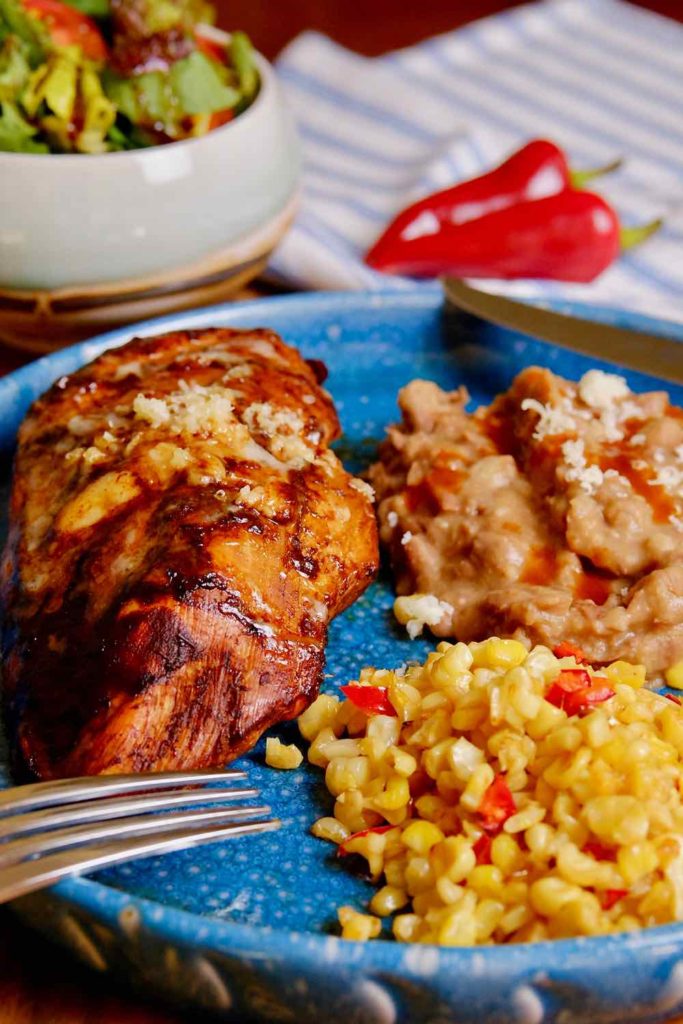 Guajillo Chili Chicken served on a blue plate with Mexican corn and Skinny Refried Beans.  Salad bowl and red chili peppers in background with white and blue striped linen.