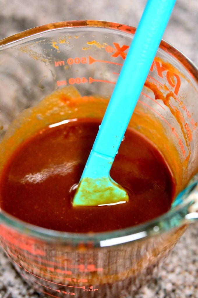 Glass pyrex measuring cup showing strained Guajillo chili sauce with blue spatula set inside cup.