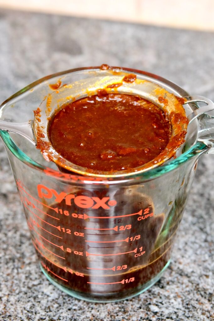 Picture depicts chili sauce in a strainer set over a glass pyrex measuring cup.