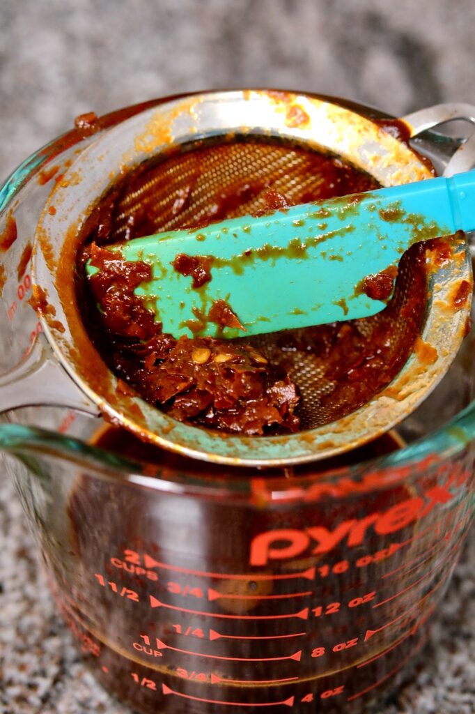 Remnants of chili sauce in strainer set over glass pyrex measuring cup, with small spatula being used to push puree through sieve.