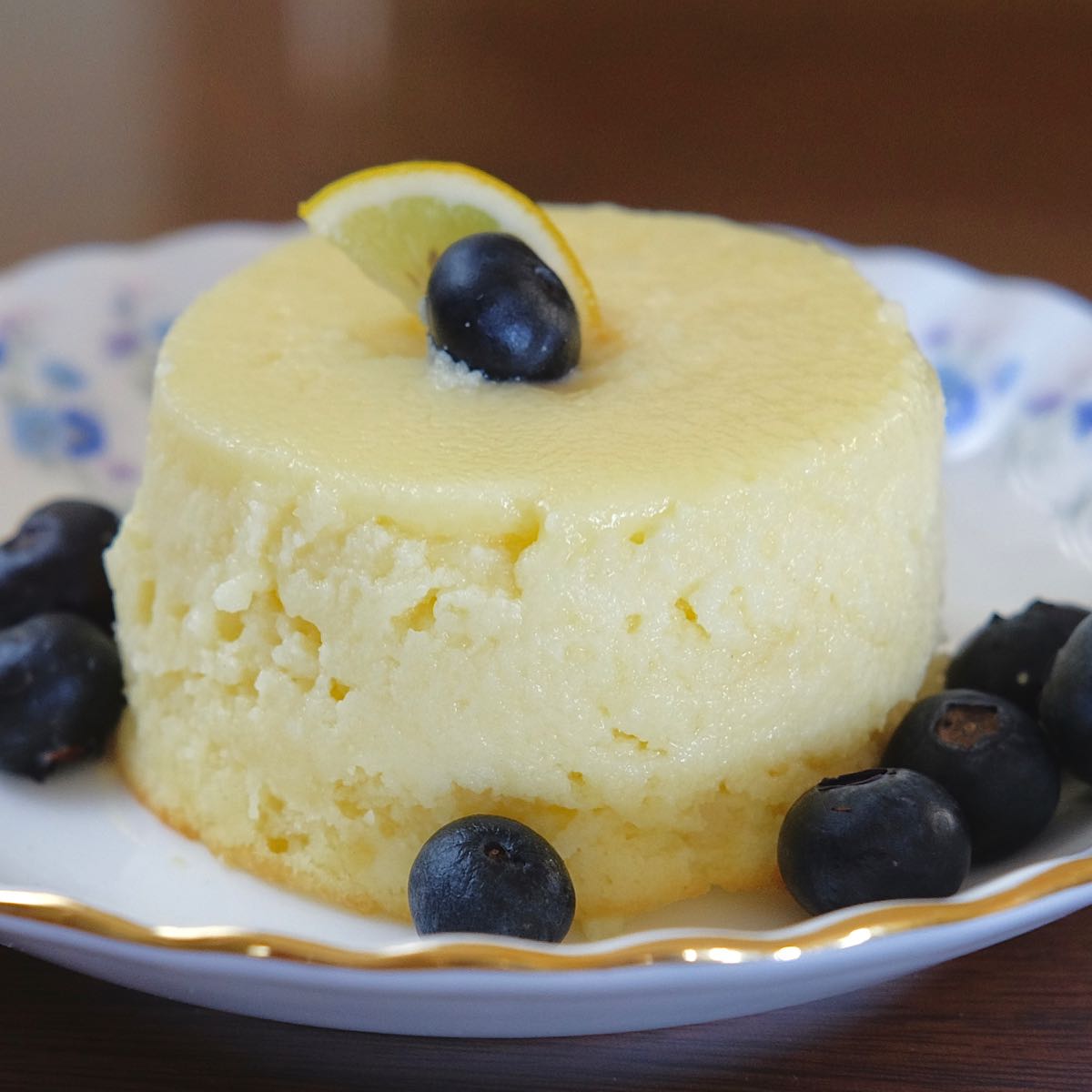 Lemon Pudding Cake on white and blue plate garnished with a thin slice. of lemon and fresh blueberries.