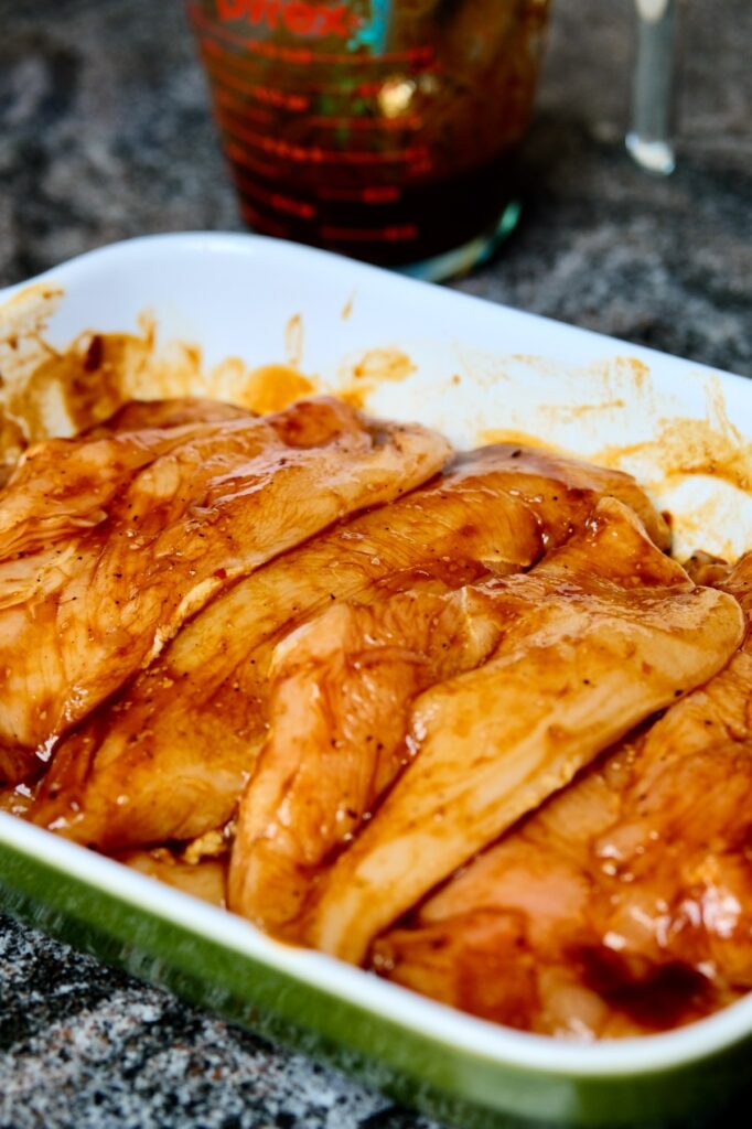 Chicken cutlets marinating in Guajillo Chili Sauce are nestled in a green and white baking dish. Glass measuring cup of remaining sauce set in background.