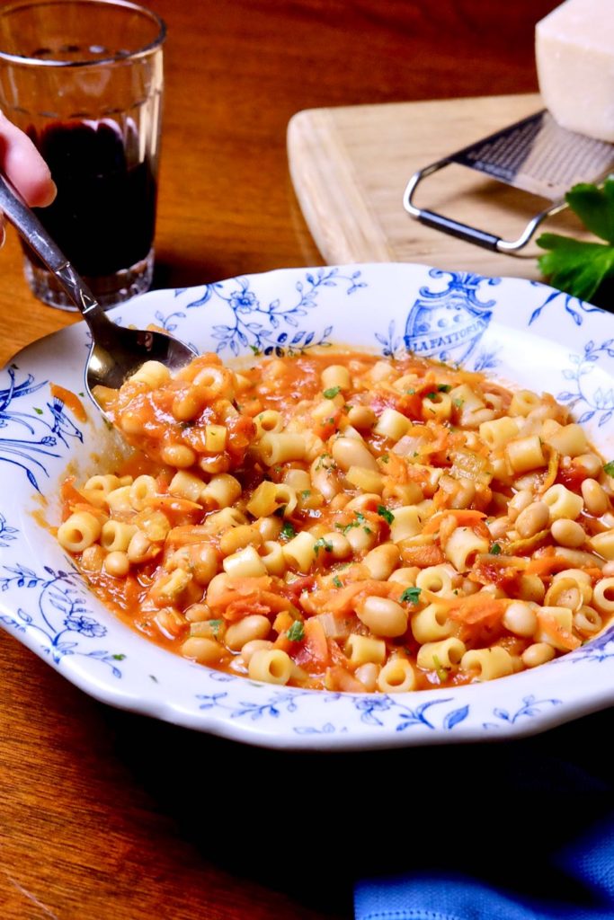 Pasta e fagioli served in a white and blue Italian bowl with spoon shot. Parmesan cheese and grater setting on board in background.