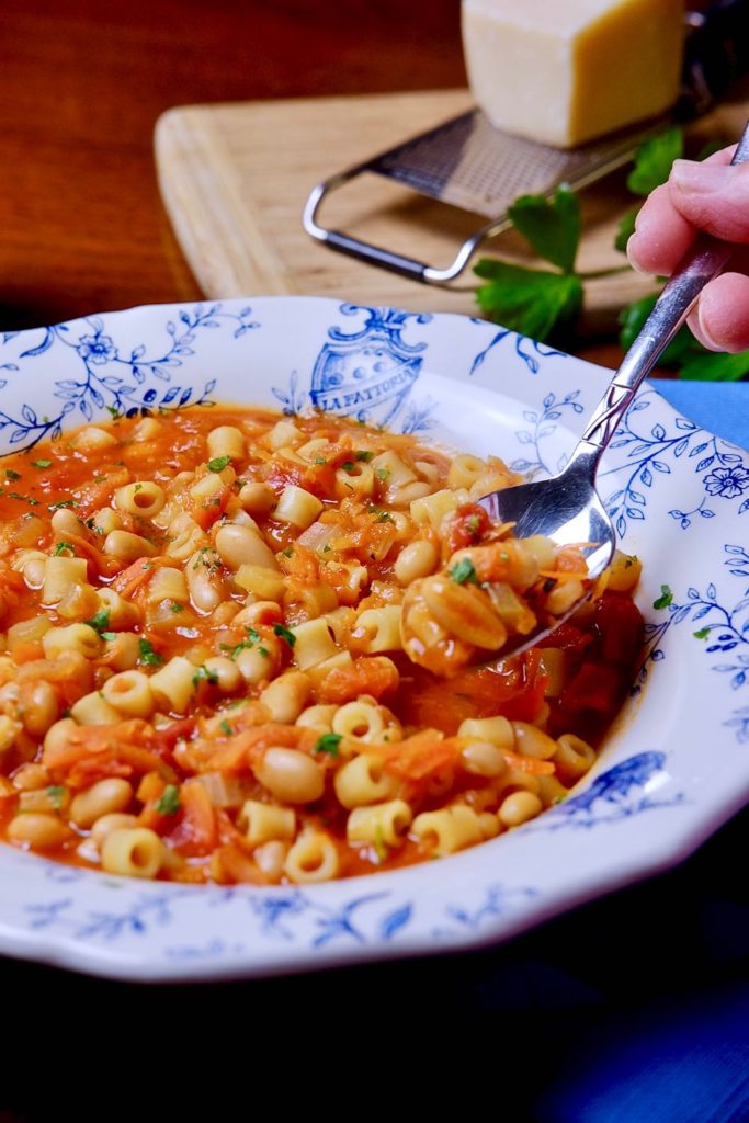 Pasta e fagioli served in a white and blue Italian bowl with spoon shot.  Glass of wine and parmesan cheese and grater in background.