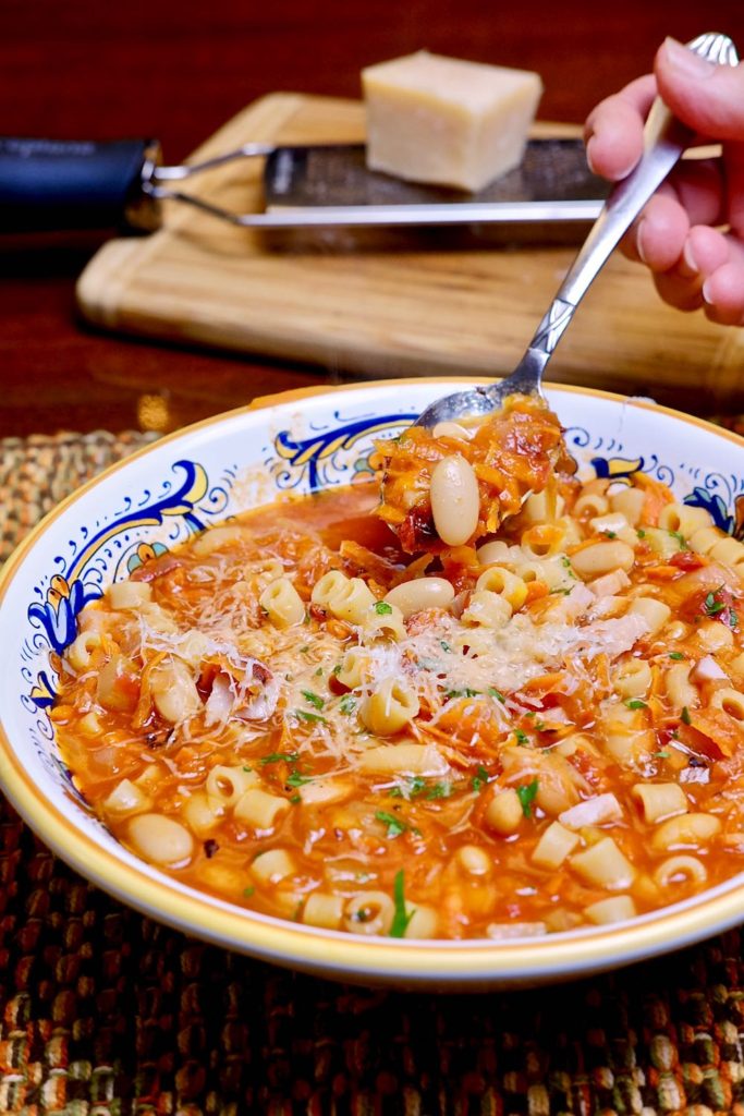 Pasta e Fagioli in blue and gold Italian bowl with bite shot of spoon.  Maple board with cheese setting on cheese grater in background.