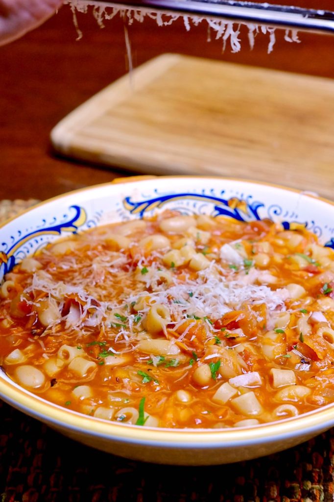 Pasta e Fagioli in blue and gold Italian soup bowl with cheese being grated over top.  Maple board in background. 