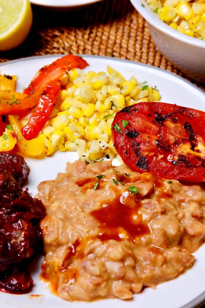 Skinny Refried Beans served on white plate with grilled tomatoes and peppers, corn, and carne adovada.